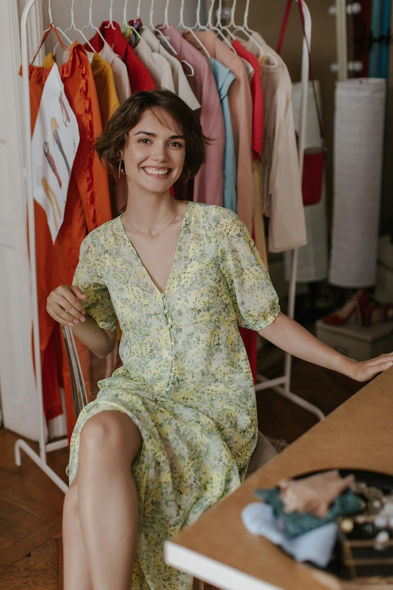 happy excited curly brunette shorthaired woman stylish floral dress smiles sincerely sits chair poses dressing room 197531 29857